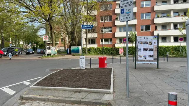 Ein Foto einer Straßenecke in einer städtischen Umgebung. Im Vordergrund befindet sich ein rechteckiger Garten, der von einem grauen Steinpflaster umgeben ist. Eine weiße Verkehrsinsel markiert eine Fußgängerüberquerung. Auf der linken Seite befindet sich ein Straßenschild mit dem Text „Stapelstraße“ und ein Schild mit der Aufschrift „3 Std“ und „Werktags 9-18 Uhr“. Im Hintergrund befinden sich mehrere mehrstöckige Gebäude. Ein roter Abfallbehälter und ein Schwarzes Schild mit Flyern stehen auf der rechten Seite.