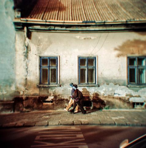 The photograph captures two people walking along an old, weathered wall with faded plaster and peeling paint. The building has a corrugated roof and several worn blue-framed windows that add a touch of symmetry to the scene. The soft focus and vignetting, characteristic of the Holga camera, create a dreamy and nostalgic atmosphere.
The colors are heavily shifted — the sky and walls glow with warm amber tones, while the shadows and windows take on surreal turquoise-blue hues. This unusual palette, produced by Lomochrome Turquoise film, transforms an ordinary street moment into something dreamlike and cinematic, as if reality itself were subtly misaligned