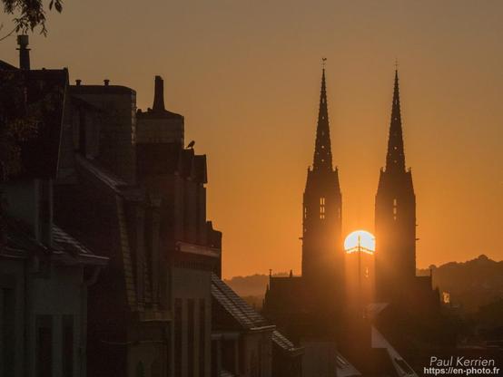 Photo en couleur à contre-jour à l'aube présentant, notamment le soleil passant juste entre les tours de cathédrale. La photographie a été prise depuis une rue en pente dont on aperçoit les toits et les cheminées à gauche du cadrage. Le ciel son nuage est de couleur dorée.