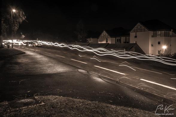 Photographie monochrome présentant les filés ondulés des lumières frontales des participant(e)s une course nocturne, passant ici sur une rue.