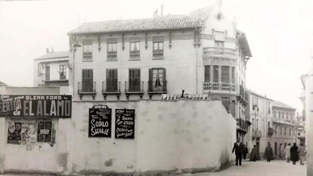Archival photo of a building group with some people walking the street beside it. The foreground is dominated by a curved whitewashed wall which is adorned with hand-painted sign boards advertising film screenings.