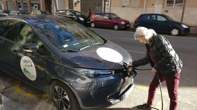 Una dona posa el cable elèctric a l'endoll del vehicle, situat a la part de davant. El vehicle està al carrer i és propietat de la cooperativa de cotxes compartits Ecotxe.