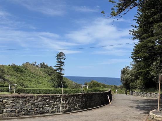 A curved stone wall along a roadway, enclosing an empty garden, surrounded by greenery with a couple of hoop pines. Blue Sky with scattered cloud above, blue sea in the distance.