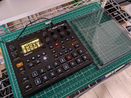 Elektron Digitakt Mk1, un aggeggio nero, di dimensioni quasi quadrate, con svariati tasti illuminati e un piccolo schermo a luce gialla.