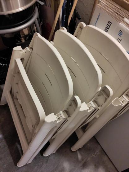 Drei zusammengefaltete weiße Kunststoffstühle in einem Kellerabteil. Drumherum etwas Krempel.