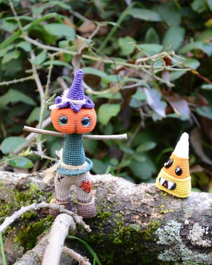 Épouvantail au crochet, avec une tête de citrouille orange et un chapeau violet, il se tient sur une branche moussue. Un peu en arrière de lui, une petite friandise au crochet orange, jaune et blanche avec des dents pointues semble le regarder.