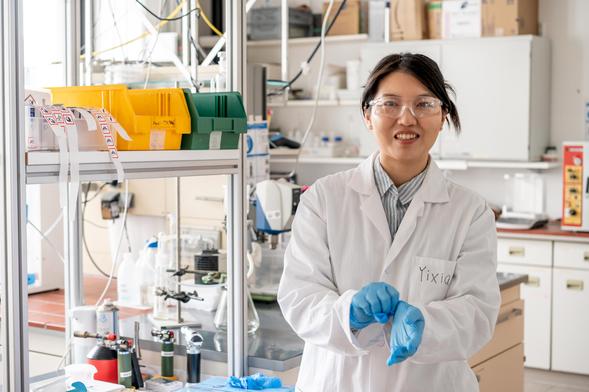 A researcher standing in her lab smiling, wearing a white lab coat and pulling off her gloves.