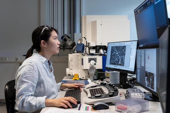 A researcher working at a computer station with multiple monitors and a microscope setup, analyzing high-resolution images of materials or samples in a laboratory environment.