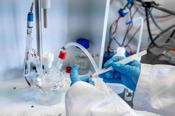 Close-up of a researcher’s gloved hands handling laboratory instruments connected to glassware and tubes. The setup appears to be part of an electrochemical or chemical experiment.
