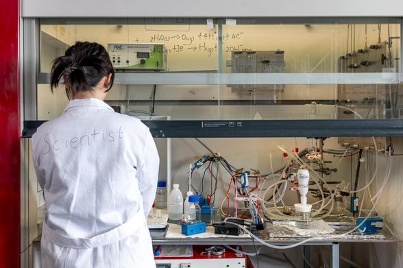 A scientist wearing a lab coat stands in front of a laboratory fume hood with experimental equipment and chemical setups. Handwritten equations are visible on the glass, and various instruments and wires are arranged inside the hood.