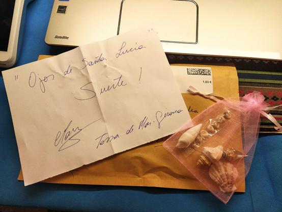 Una carta con el texto "Ojos de Santa Lucía Suerte!" con una firma y la mención a Tossa del Mar (Girona). También incluye una bolsita con conchas recogidas en la playa, entre ellas, precisamente, ojos de Santa Lucía.