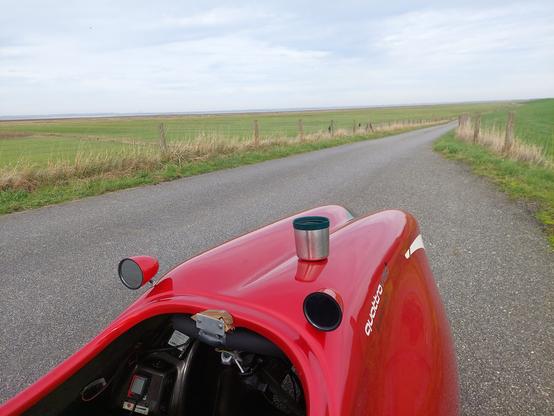 Mein Velomobil steht auf einem Asphalt-Weg auf dem Deich. Auf der Nase steht eine Tee-Tasse. Im Hintergrund absolut flache Feuchtwiesen. Am Horizont sieht man das gegenüberliegende Ufer des Jadebusen.