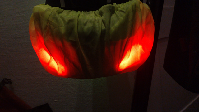 Ein im Dunkeln an einem Sattel hängender Lumos Ultra Fahrradhelm mit einem dünnen, gelben Regenschutz übergestülpt. Der Helm hat zwei schräge Rückleuchten, die angeschaltet sind und den Regenüberzug aussehen lassen wie zwei böse drei blickende, schlitzförmige Augen eines Außerirdischen.