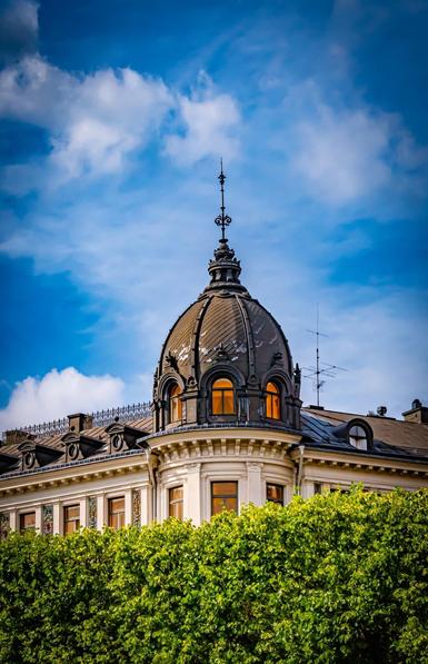 Ornate building with a dark domed roof featuring decorative elements and circular windows. Warm lighting from the windows suggests interior illumination. Lush green foliage partially obscures the lower part of the structure. The sky is blue with scattered clouds.
