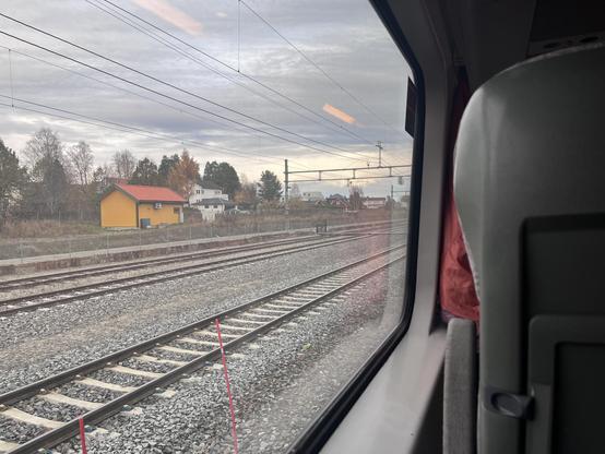 View through a train window. You can spot train tracks and a yellow house. To the right you can see a train seat.