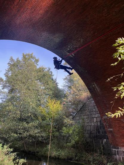 Das Bild zeigt eine Person, die sich von einer alten Steinbogenbrücke abseilt. Die Person trägt einen Gurt und ist mit einem Seil gesichert. Im Hintergrund sind Bäume und ein Fluss zu sehen. Der Himmel ist blau.
