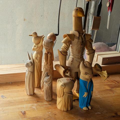 Group of woodcarvings on a workbench. A bat inspired creature, a pipistrelle bat, a dragon with a duck on its head, a mantis humanoid creature with a long skirt, a humanoid cuttlefish wearing a dress, a goode knight with shield and a mace where sits a red robin, and humanoid creature with 4 eyes in a blue dress