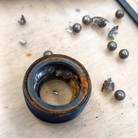 Au premier plan, posé sur une surface beige, un objet métallique cylindrique rouillé aussi bien à l'intérieur qu'à l'extérieur avec bouts de métal qui se détachent de la partie interne.
À côté, en haut à droite de l'image sont éparpillés des bouts de métal, certains rouillés, et des billes métalliques.
C'est une cuvette de roulement d'un boîtier de pédalier de vélo, tellement usée et rouillée que des morceaux ont cassé dedans.