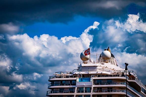 Large cruise ship deck with multiple levels, featuring a Norwegian flag and two dome-shaped structures on top. The background consists of a dramatic sky with dark, dense clouds and patches of blue.