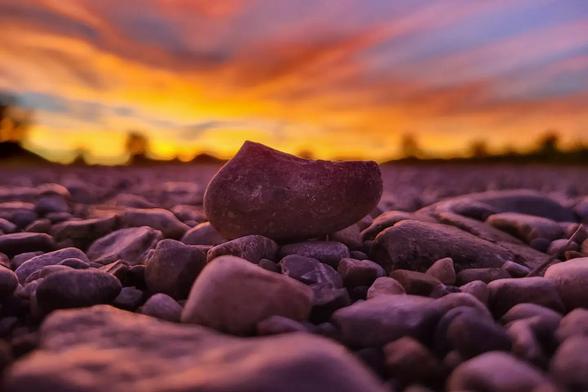 A close-up of small stones on the ground with a single larger rock in focus, set against a vivid orange and purple sunset sky.