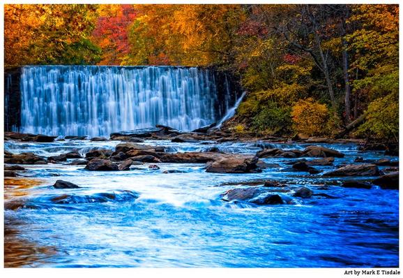 Color photograph in horizontal format of a fall landscape with a waterfall at the end of a large creek. Both sides of the water are framed with fall foliage. The waterfall in the distance is old but manmade.