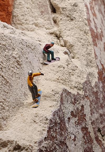 Miniaturfotografie: Zwei Snowboarder fahren hangabwärts. Inszeniert an einer Zimmerwand, wo der Wandputz linear abgebrochen ist