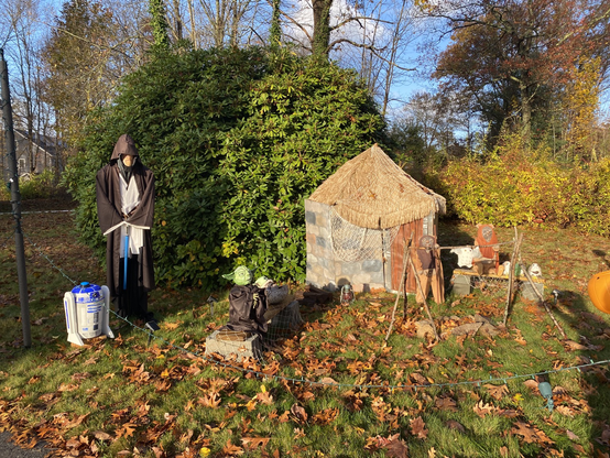 Star Wars themed Halloween decorations beside a bush in a leaf-covered yard. Left to right: R2D2, Obi Wan Kenobi, Yoda, Baby Yoda, a hut and two ewoks