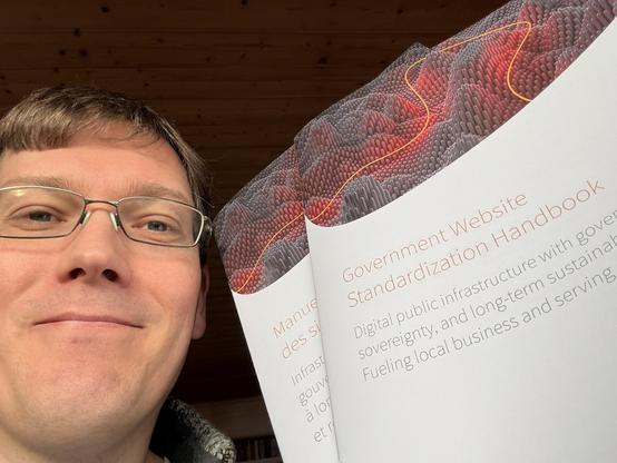 A person wearing glasses smiles while holding up two copies of the Government Website Standardization Handbook. The covers feature a textured red and black design with orange lines.