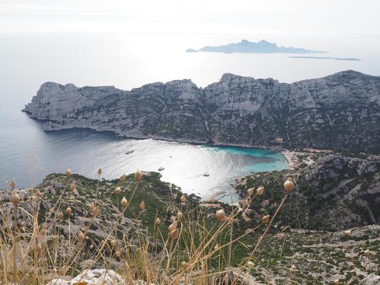 Sous un ciel un peu voilé, une barre rocheuse s'avance dans la mer Méditerranée. Devant, la vue est prise depuis un sommet entre roches et herbe rase. On voit trois petits bateaux dans l'eau bleue foncée, tandis que l'eau au bord est bleue clair.  Au tout premier plan, quelques herbes hautes et jaunes. Derrière la barre, 2 petites îles. Ici la mer est presque blanche et éblouissante.