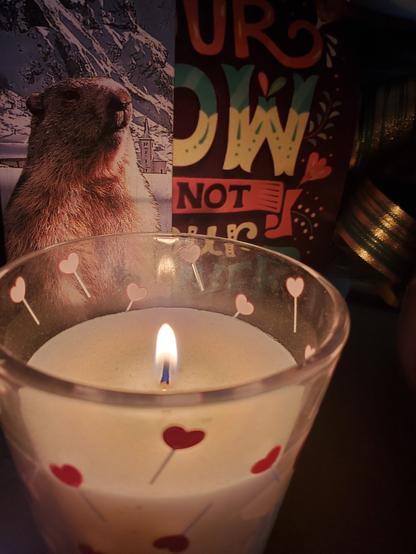 Une bougie allumée dans un verre couvert de sucettes coeur rose devant une affiche,  un nœud de coussin de Lyon et un marque page marmotte