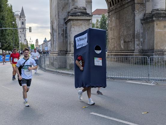 Un homme court le marathon à l'intérieur d'une boîte qui ressemble à une penderie en tissu, des trous circulaires. Laisse passer les bras et un trou  au niveau de la tête lui permet de voir où il va. Il porte la boite avec ses mains. Sur le coté, au-dessus du trou circulaire, est inscrit : A book can change a life