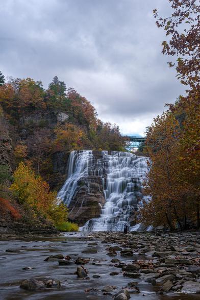 Ithaca Falls cascading 150 feet over layered shale rock formations, photographed from creek level with rocky streambed in foreground and autumn foliage along gorge rim under overcast sky