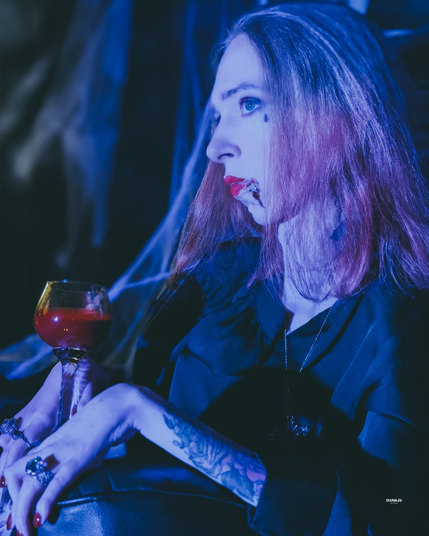 Seitliches Porträt von mir mit langem, leicht roséfarbenem und blondem Haar in kaltem Blau-Licht. Rotes, glänzendes Lippen-Make-up mit etwas „Blut“-Effekt am Mundwinkel. Ein schwarzes Outfit, große Ringe und ein tätowierter Unterarm sind sichtbar. In den Händen ruht ein Kelchglas mit dunkelroter Flüssigkeit. In meinem Blick liegt keine Hast, kein Zucken im Kiefer, es ist die elegante Geduld eines Wesens, dessen Zeit unendlich ist. Das Blaulicht küsst die Iris und zeichnet Wange und Nase wie Marmor: fern, wach, unnahbar. Im Hintergrund eine dunkle Bühne mit Spinnweben, kontrastreich beleuchtet.