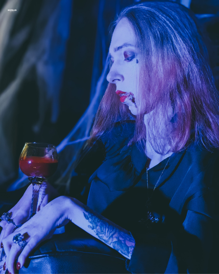 Fast die gleiche Szene aus etwas größerer Distanz, der Kopf ist leicht gesenkt. Der Blick ruht nachdenklich und verträumt, wie ein stilles Gebet, auf dem Kelch voller Blut. Mit halb geschlossenen Lidern, als lausche ich dem Flüstern der Nacht. Das blaue Licht streicht kühl über Wange und Hals.