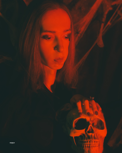 Ein frontaleres Porträt in intensivem Rotlicht. Ich blicke nach unten; die Hand liegt auf einem Totenschädel. Im Hintergrund sind Spinnweben, starke Hell-Dunkel-Kontraste erzeugen eine dramatische Atmosphäre wie bei einem Ritual.

Im Rot der Stunde senkt sich der Blick wie ein Vorhang.
Die Lider halten die Stille, die Lippen bewahren das Geheimnis.
Unter der Hand, kalter Knochen, ein stiller Chronist der Nacht.
Das Licht zeichnet scharfe Kanten an Wange und Stirn;
nichts atmet, als lausche die Szene auf einen Herzschlag.