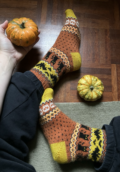 Jalat, joissa Halloween-kuvioiset kirjoneulesukat. Lisäksi kaksi pientä koristekurpitsaa, toinen lattialla ja toinen jalkoihin nojaavassa, ojennetussa kädessä; to