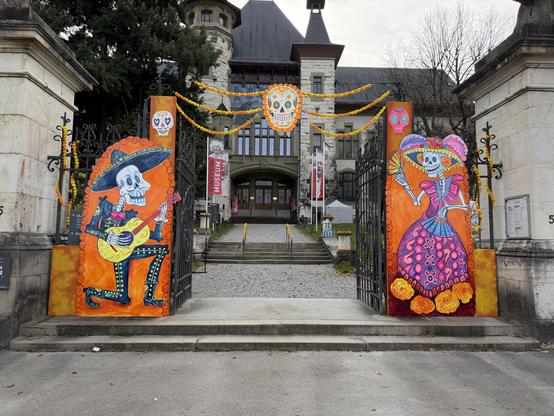 Der Haupteingang vom Museum, links und rechts tanzende und musizierende Skelette, oben von links nach rechts bunte Blumen und einen farbigen Totenkopf über den Eingang 
The main entrance to the museum, with dancing and musical skeletons to the left and right, colorful flowers above from left to right, and a colorful skull above the entrance