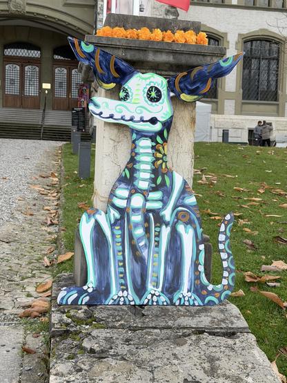 Ein Hund als buntes Skelett bemalt, sitzt vor eine Säule die mit bunten Blumen geschmückt ist
A dog painted as a colorful skeleton sits in front of a column decorated with colorful flowers