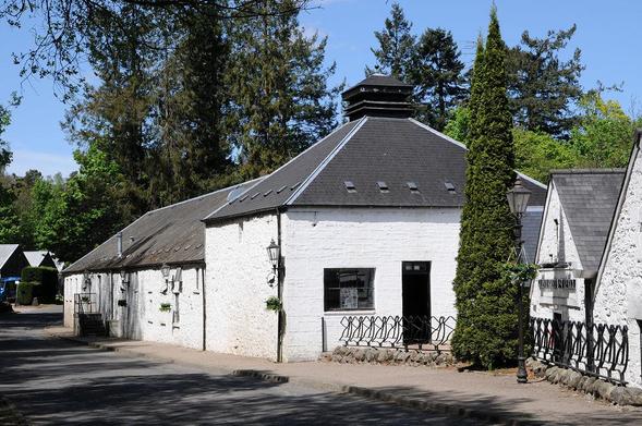 Glenturret Distillery. The image shows a road in the bottom left of the frame with a row of single-storey white buildings along its far, right side. There are two gable ends on the right and then a square building with a grey slate roof topped off with a distillery pagoda, then a run of further buildings. There are trees in the background and the sky is blue.