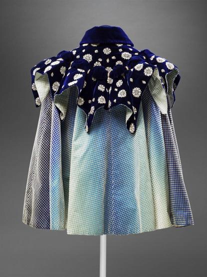 Short cloak of eight panels of velvet, with white cut pile spots on a satin ground shaded from pale to dark blue. Gathered cape of dark blue standing out in ten points, each of which is headed by a bow, with applied lace motifs. Semi-stiff standing collar of plain dark blue velvet, fastening with black hooks in front. Striped satin lining.