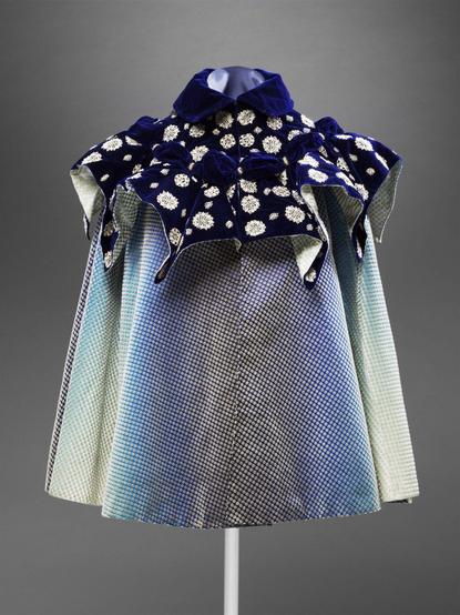 Short cloak of eight panels of velvet, with white cut pile spots on a satin ground shaded from pale to dark blue. Gathered cape of dark blue standing out in ten points, each of which is headed by a bow, with applied lace motifs. Semi-stiff standing collar of plain dark blue velvet, fastening with black hooks in front. Striped satin lining.