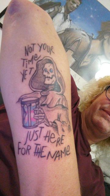 Selfie où on voit principalement mon avant bras avec un tatouage de La Mort (personnage de l'univers du Disque Monde, squelette encapuchonné avec les yeux bleus) qui tient un sablier aux couleurs du drapeau trans (bandes bleu, rose, blanc, rose, bleu) avec comme texte en majuscules (parce que La Mort s'exprime toujours en majuscules) : NOT YOUR TIME YET, JUST HERE FOR THE NAME, ce qui signifie : TON HEURE N'EST PAS ENCORE VENUE, JE SUIS JUSTE ICI POUR LE NOM.
En arrière plan, mon visage est coupé mais on me voit avec mes cheveux blonds et mes lunettes devant une affiche représentant un homme et une femme dessinés par Enki Bilal