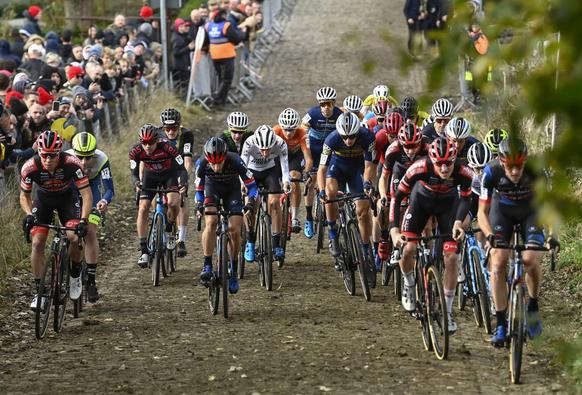 A large group of cyclo-cross racers racing towards the camera up the cobbled Koppenberg climb. On the right, spectators line the roadside behind a wire fence.