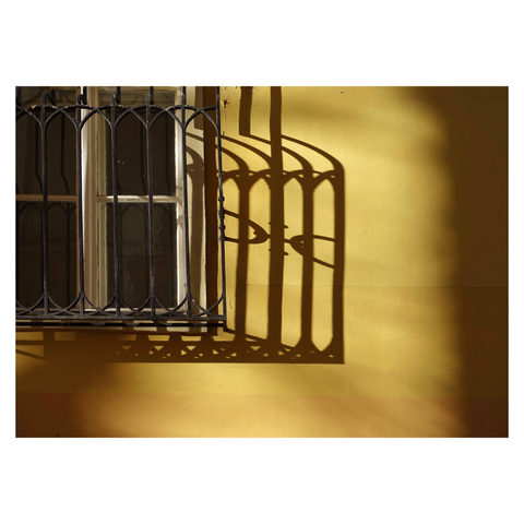 This isn't a landscape format colour image of athr corner of a white wooden window frame with an ornate metalwork surround set against a bright yellow wall. Set to the top right corner of the frame, and with light falling from the left, a strong shadow of the metalwork sits in the centre of the composition. Taken in Budapest, Hungary.
