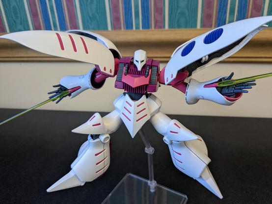 The HG Qubeley in a bizarre hovering pose on a flight stand. All its limbs are at a strange but realistic angle, and its shoulder parts are spread open. It is looking directly at the viewer, and its eyes appear to glow.