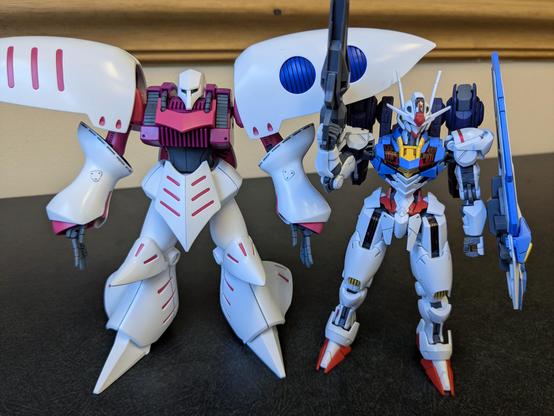 The HG Qubeley standing beside the HG Gundam Aerial. The top of Qubeley's head come above that of Aerial's, but below the tips of Aerial's antennae. The tops of Qubeley's shoulders come above Aerial entirely.