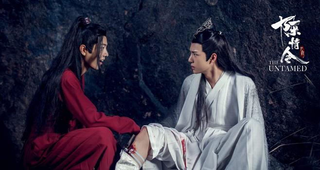 In the Xuanwu's cave, Wei Wuxian in his red underrobe crouches in front of a seated Lan Wangji who has his right trouser leg pulled up to reveal a bleeding wound just below his knee. Wuxian is speaking with a grin on his face while Wangji looks back at him with an incredulous expression.