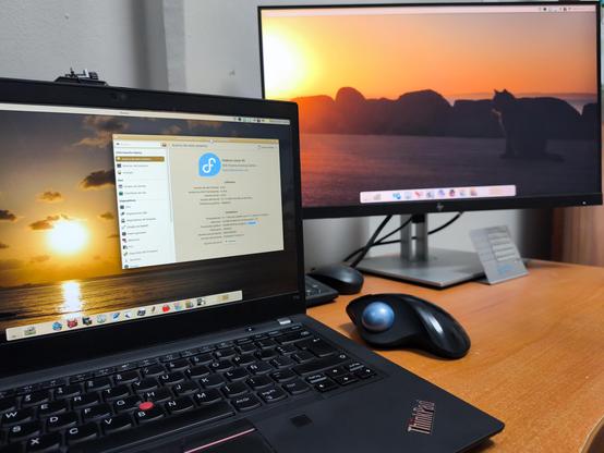 Foto de escritorio con un portátil y un monitor externo, colocados sobre una superficie de madera. El portátil, una laptop ThinkPad de color negro, está abierto y su pantalla muestra una imagen de un atardecer con el sol reflejándose en el agua. A su lado, un monitor de pantalla ancha también exhibe la misma imagen del atardecer, creando una vista de doble pantalla.
Sobre el escritorio, frente al monitor, se encuentra un ratón ergonómico de color negro. La pantalla de la laptop muestra la ventana del centro de información de KDE Plasma, indicando que el sistema es Fedora 43. Los fondos de pantalla de ambos monitores son fotos del amanecer visto desde la orilla del mar.