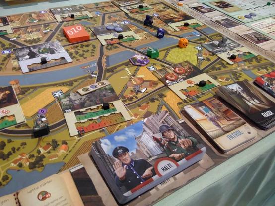 Eine Nahaufnahme vom Brettspiel „Dutch Resistance: Orange Shall Overcome!“, schräg von oben fotografiert. Der Spiellan zeigt eine Stadt von oben. Darauf verteilt liegen mehrere Pappmarker sowie Holzfiguren. Vorne sind mehrere Kartenstapel, u.a. einer mit einem Grenzsoldaty an einer rot-weißen Schranke mit einem „Halt“-Schild.