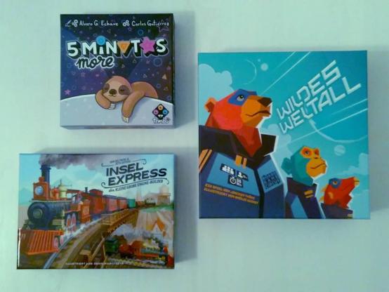 Drei Spielecover, von oben fotografiert: „5 minutes more“, mit einem illustrierten, schlafenden Faultier. „Wildes Weltall“ mit drei anthropomorphen, in Astronautenkleidung (ohne Helm) nebeneinander stehenden Tiere, die ernst nach vorne, leicht nach oben in den Himmel schauen, gezeichnet in einem eher eckigen Stil. „Insel-Express: Der kleine große Engine-Builder“ mit einer Dampflok mit vielen Waggons auf einer Holzbrücke im Vordergrund, unter der Brücke am unteren Rand fährt weiter hinten in kleiner ein weiterer Dampfzug.